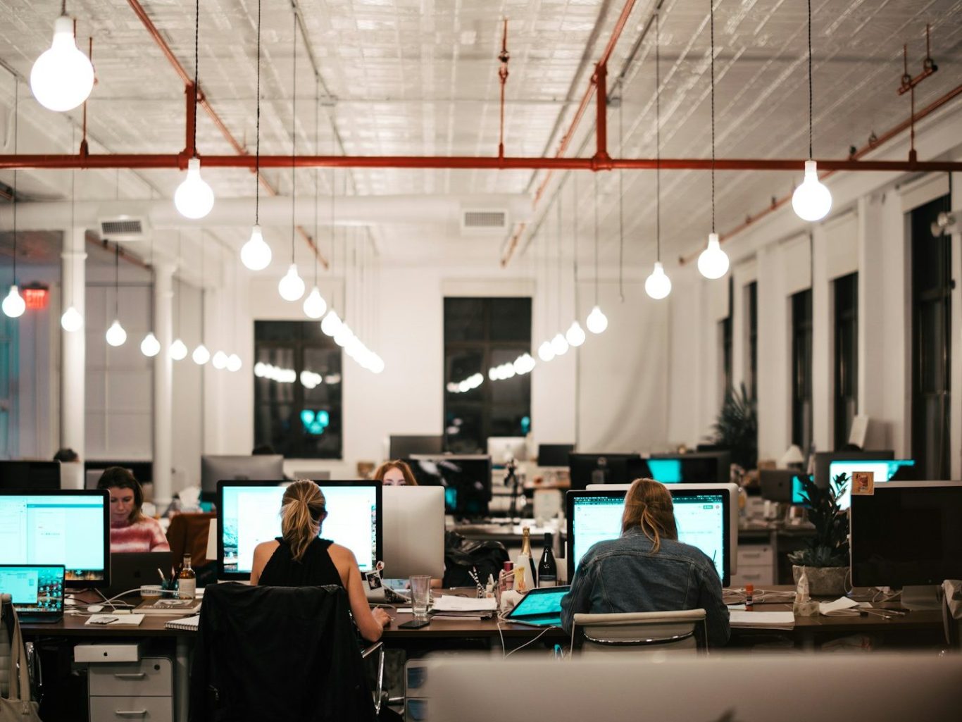 Bureau moderne avec plusieurs personnes travaillant sur des ordinateurs, éclairage suspendu.