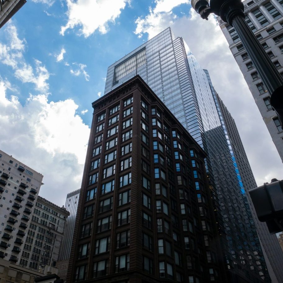 Immeuble moderne en verre sur fond de ciel nuageux, entouré de bâtiments plus anciens.