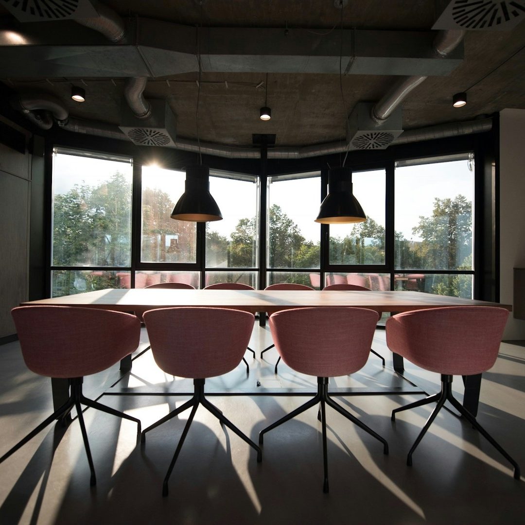 Salle de réunion moderne avec une table et des chaises roses, grandes fenêtres, éclairage pendentif.