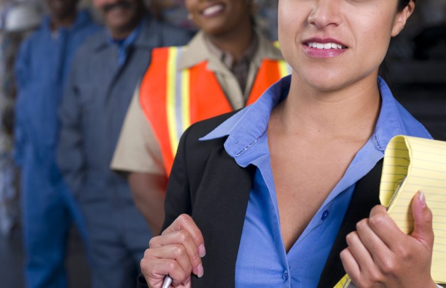 Femme en costume avec un carnet, entourée d'hommes en uniformes de travail.