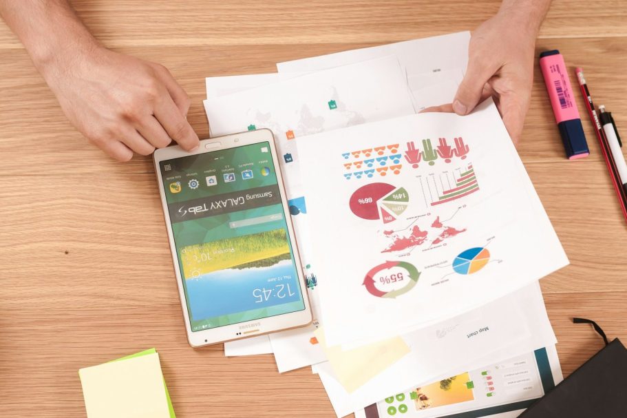 Main d'une personne tenant une tablette, entourée de graphiques et de documents sur une table.