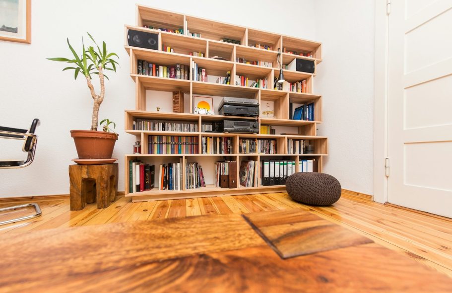 Bibliothèque en bois avec des livres, plante, pouf et chaise dans une pièce lumineuse.