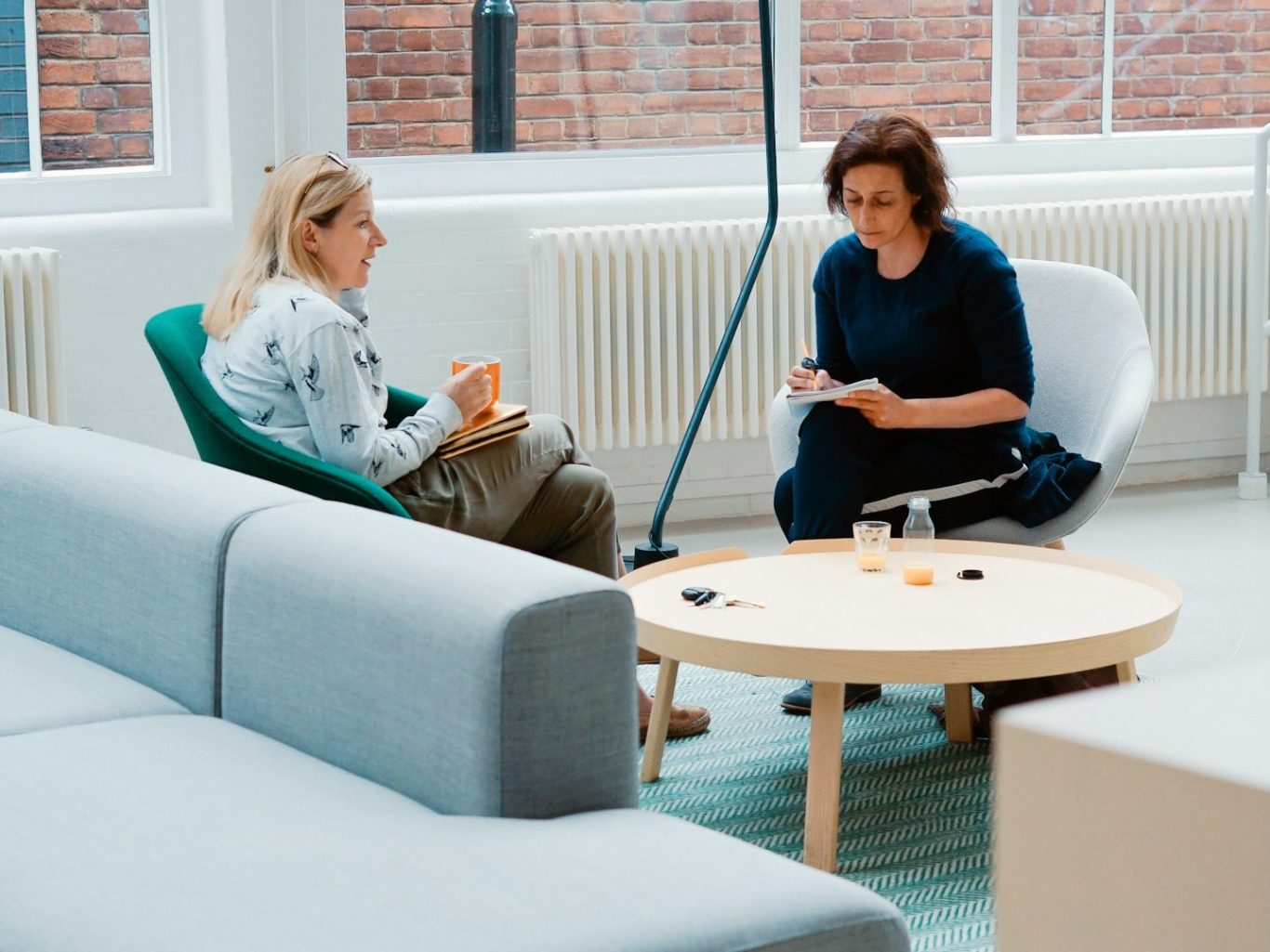 Deux femmes discutent assises sur des fauteuils autour d'une table basse dans un intérieur lumineux.